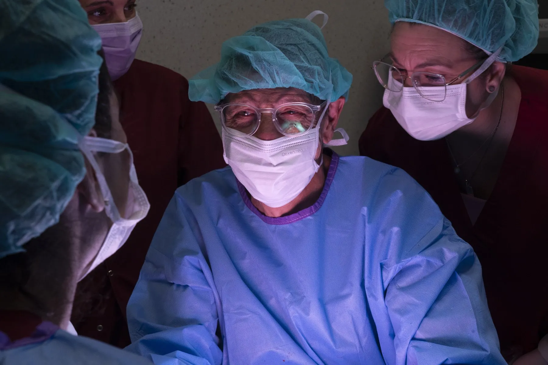 A surgeon in a mask and glasses watches the operation, surrounded by his team