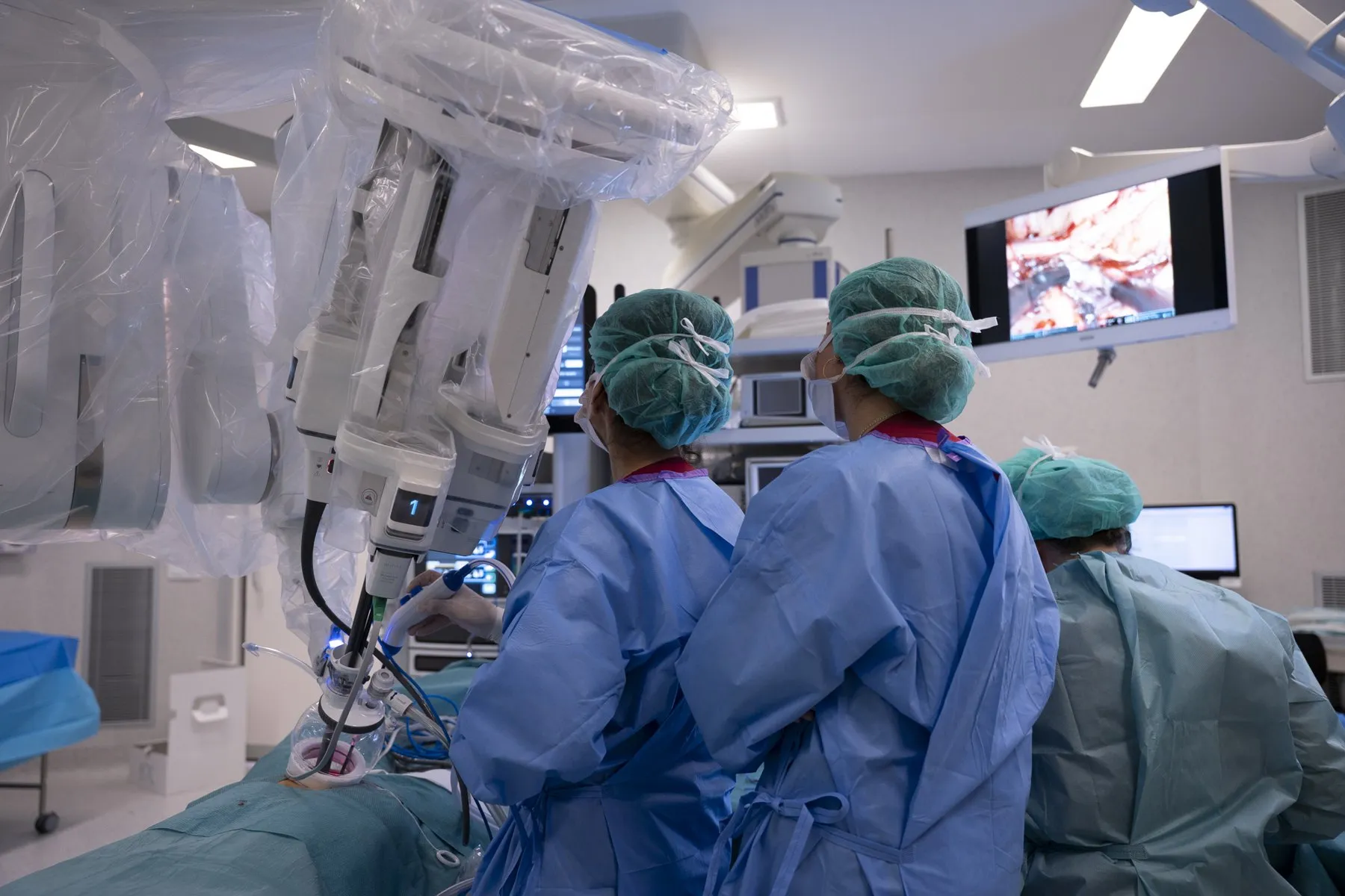 A surgical team in blue caps and gowns next to the arms of a surgical robot in an operating theatre