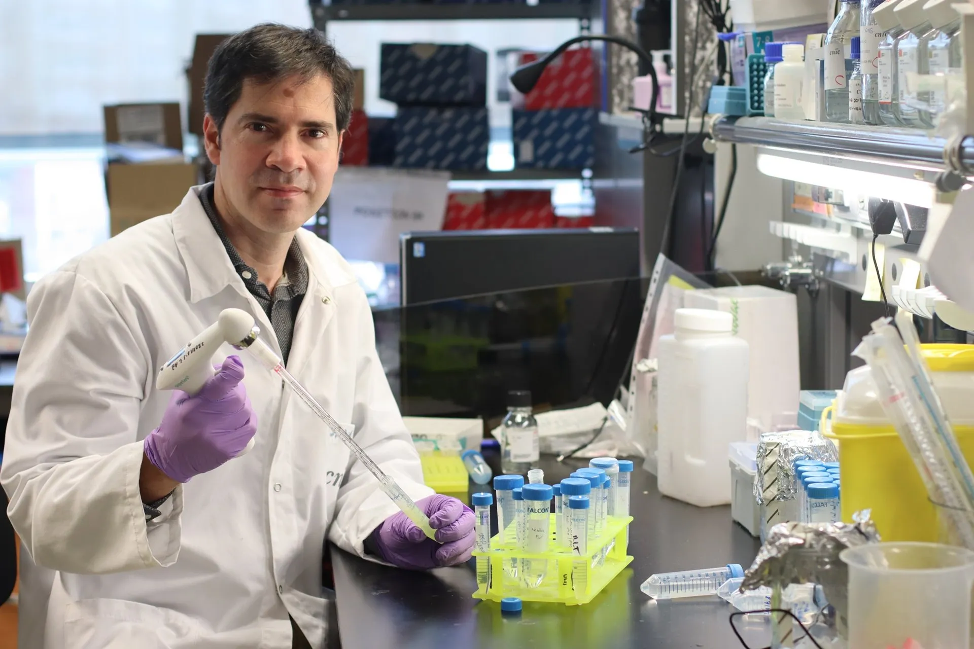David Sancho, in a white lab coat and purple gloves, holds a pipette next to a yellow rack of test tubes in his lab