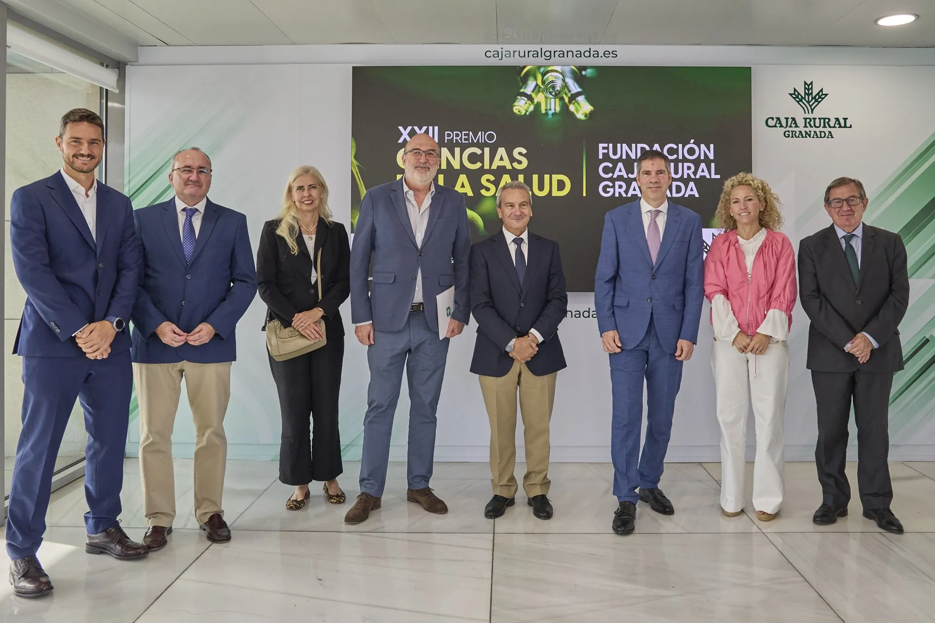 Eight people stand in front of a screen reading "XXII Premio Ciencias de la Salud Fundación Caja Rural Granada"; David Sancho, in a blue suit and pink tie, is sixth from the left