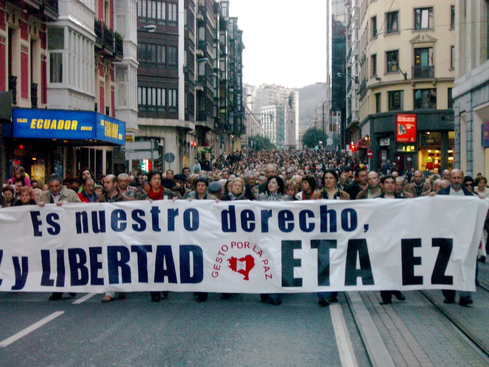 The front of a march in a Bilbao street, with a large white banner ending in the words "ETA EZ" and the Gesto por la Paz logo