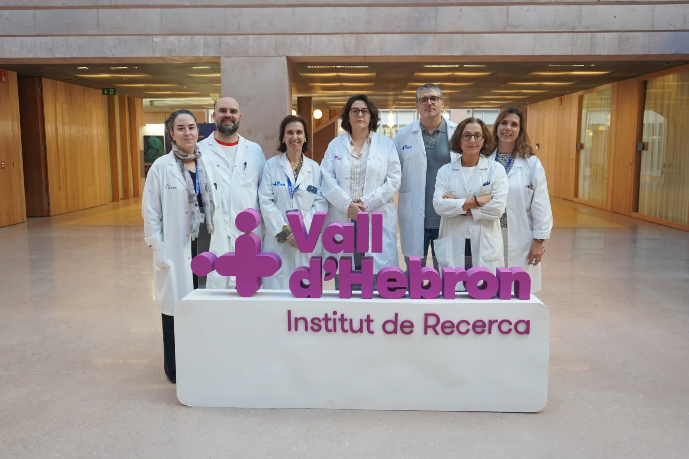 Seven people in white lab coats stand behind the purple Vall d'Hebron Institut de Recerca sign in the building's lobby