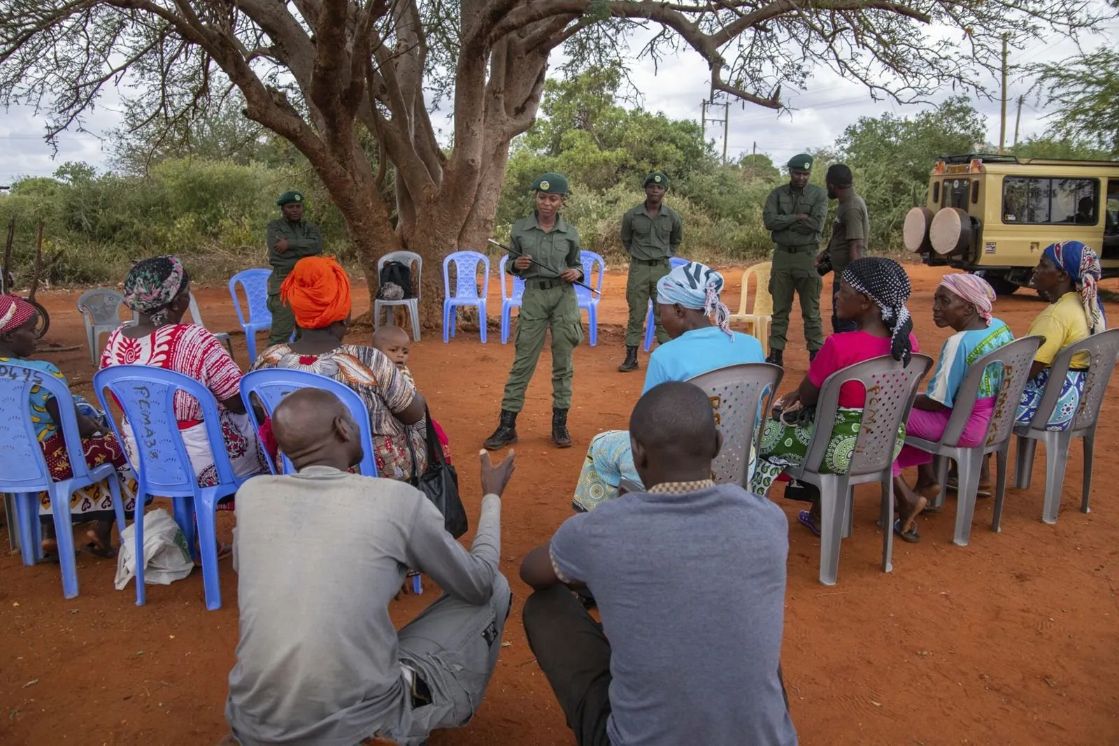 Residents of Kiteghe village sit in a circle under a tree, listening to a uniformed ranger
