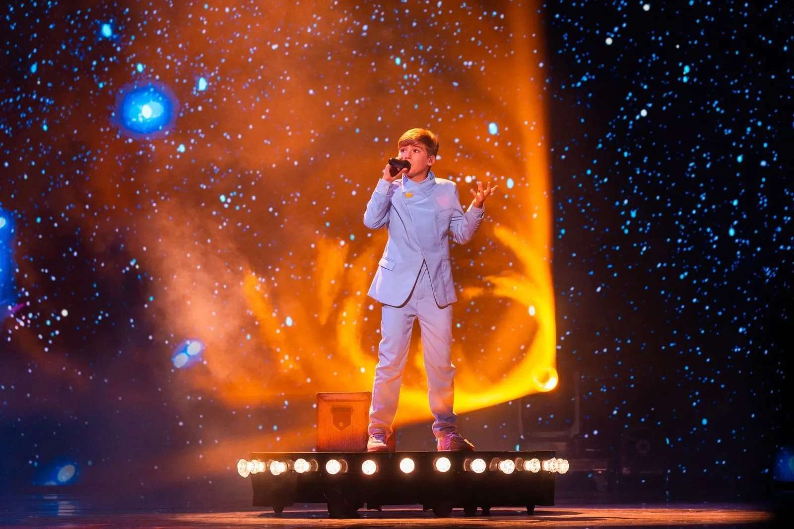 Lucas Paulano stands on a platform lined with spotlights, singing in front of a screen showing a starry sky and an orange trail