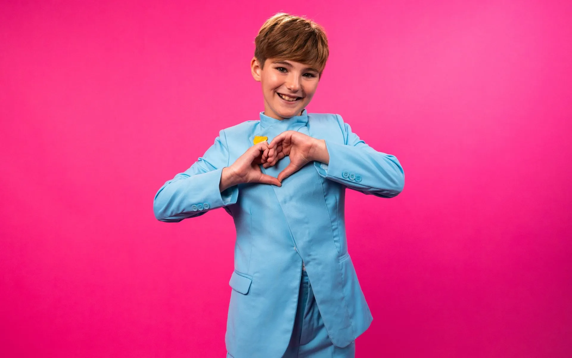Lucas Paulano, smiling, makes a heart shape with his hands against a pink background