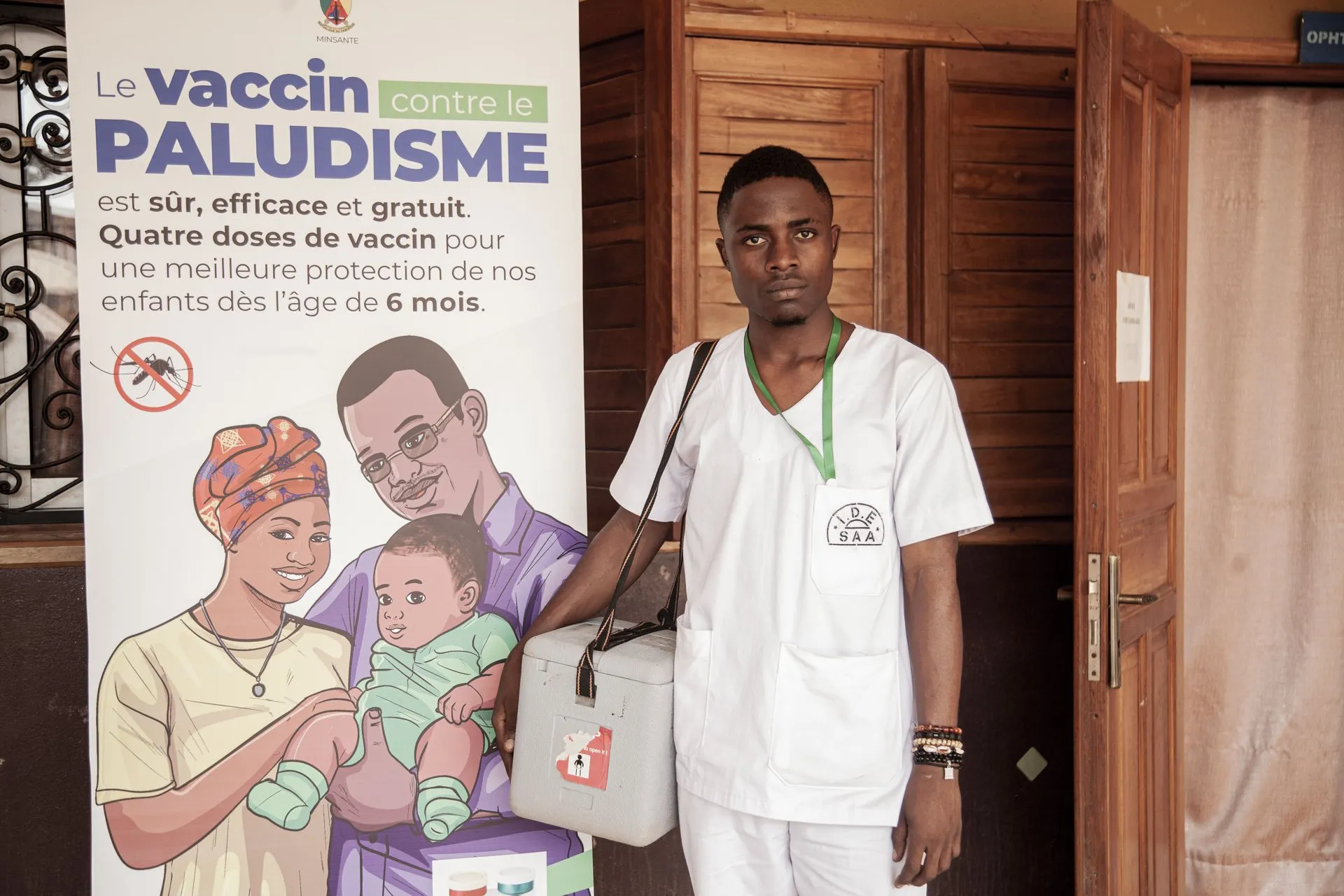 A vaccinator holding a cool box stands next to a poster saying the malaria vaccine is safe, effective and free