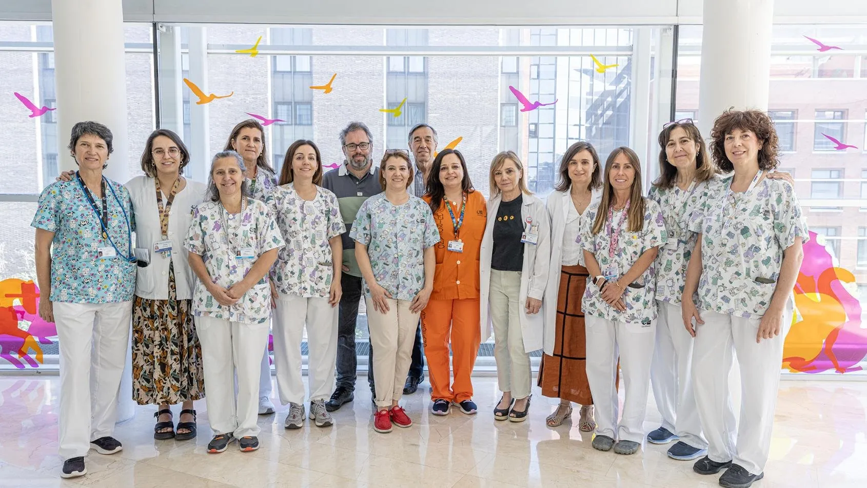 Fifteen health workers, most in children's print scrubs or white coats, pose together in a glass-walled hallway decorated with colourful bird silhouettes