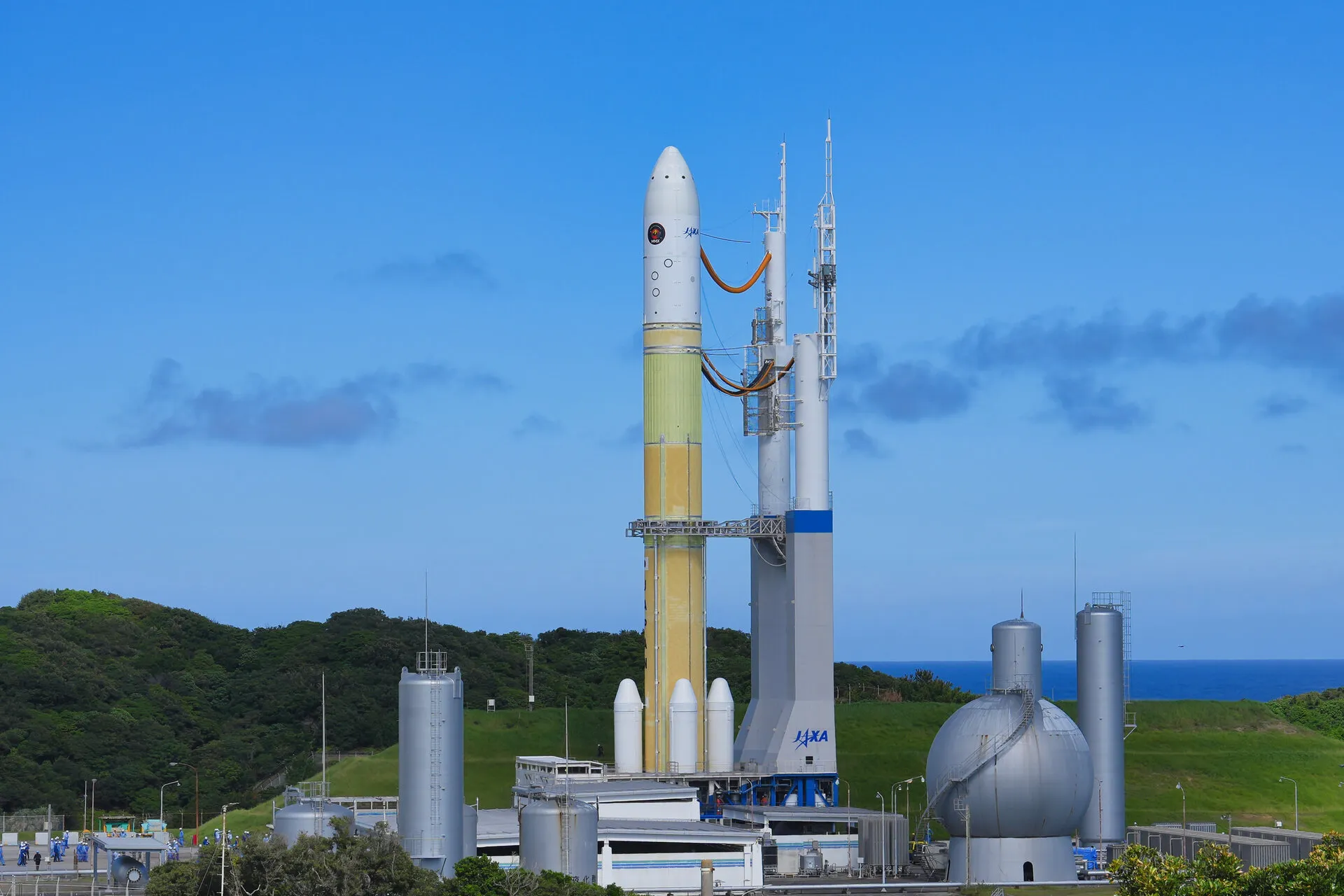 The white-and-yellow H3 rocket carrying the MMX probe stands on its mobile launch platform beside the umbilical tower at Tanegashima, with the sea behind