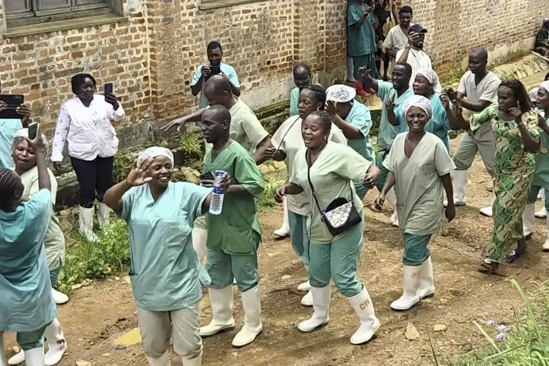 Health workers in green scrubs and white boots dance and sing on a dirt path beside a brick building, while others film them on their phones