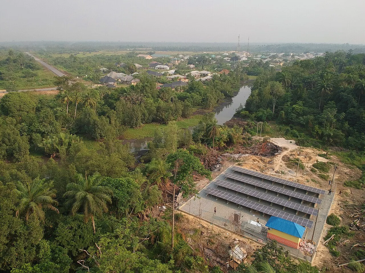 Aerial view of a solar panel plant next to a village surrounded by palm trees and forest