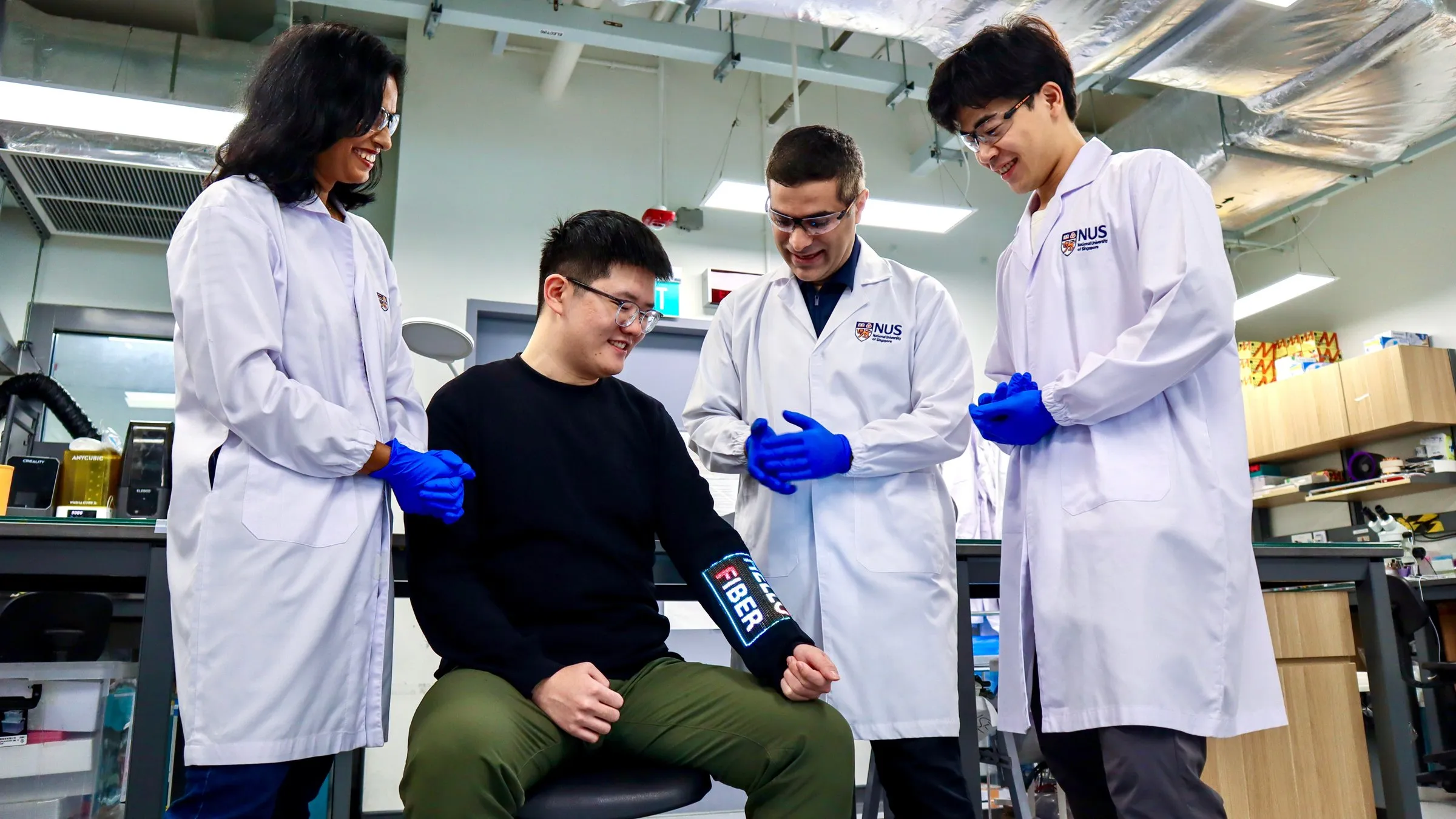 Four researchers in lab coats smile as one of them, seated, wears the sleeve on his forearm with the word FIBER lit up