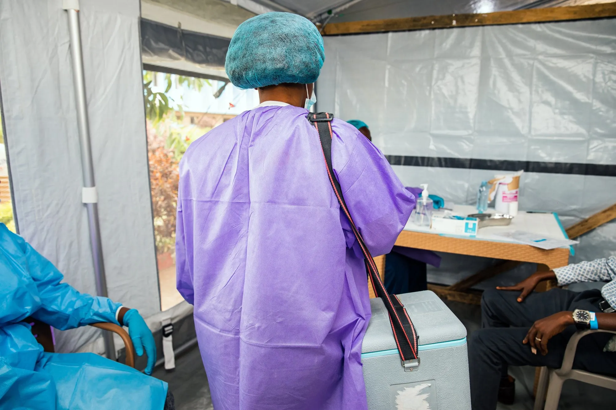 A vaccinator in a purple gown and hairnet, with a cool box over her shoulder, walks into a tent where several people sit waiting