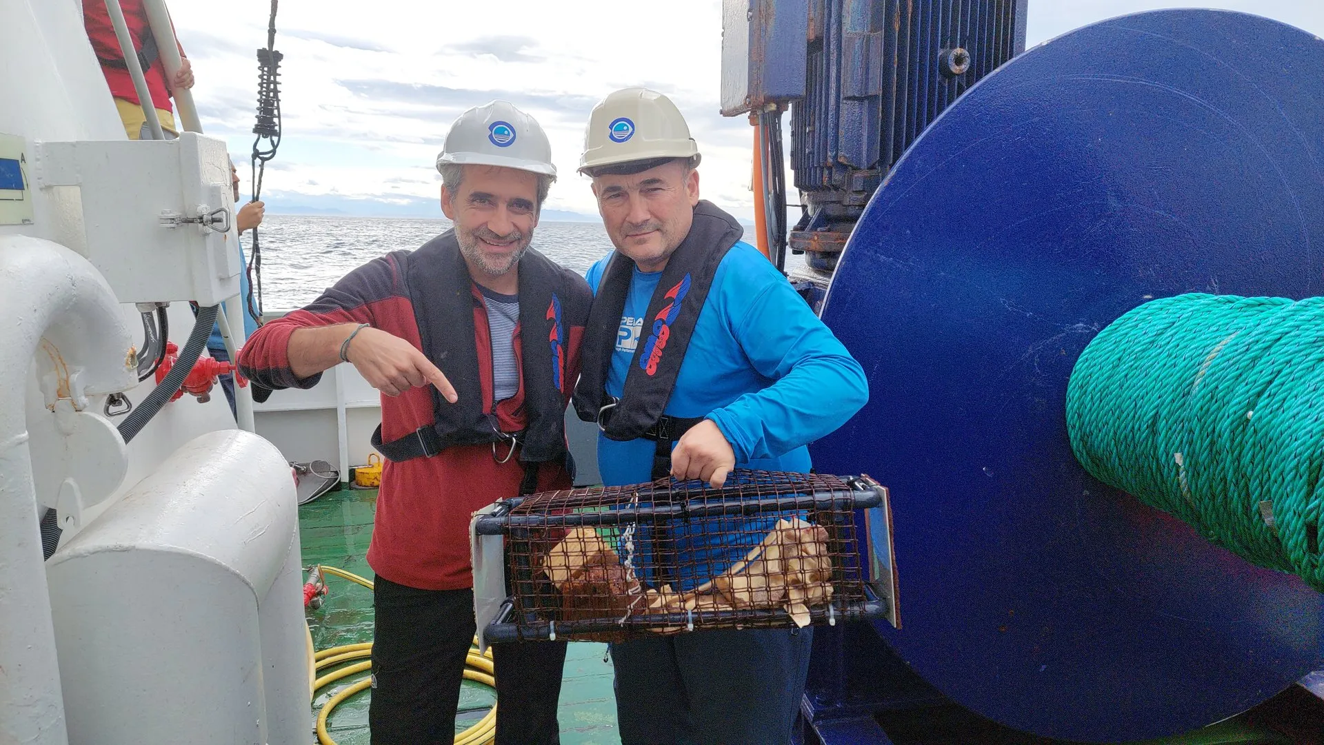 Two researchers in hard hats and life jackets smile on a ship's deck next to a metal basket full of bones