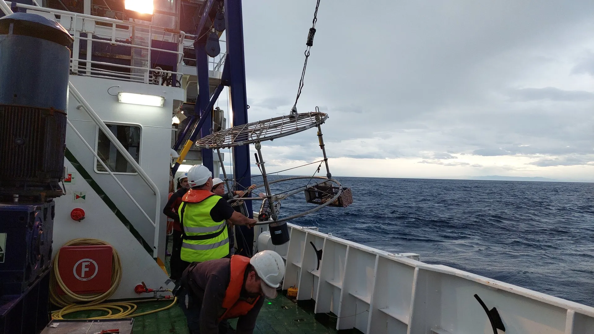 A metal frame with floats hanging from a crane over a ship's deck, with the sea behind