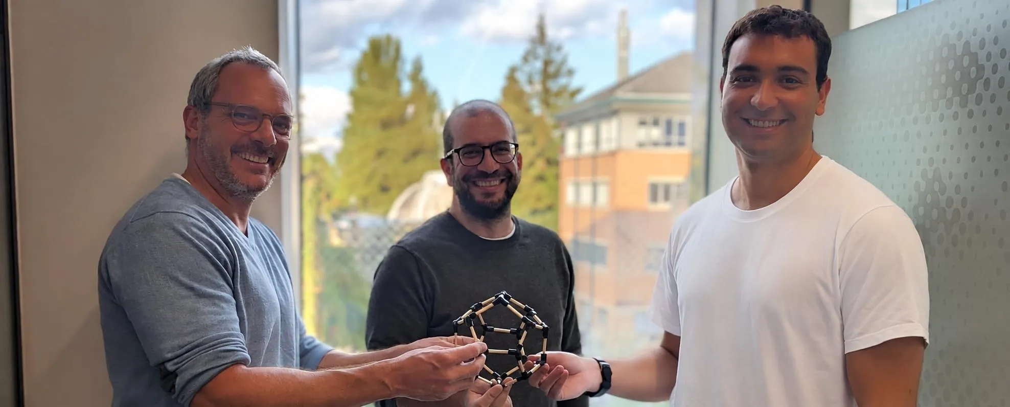 Three smiling researchers hold a molecular model made of dark pieces joined into pentagons, next to a window