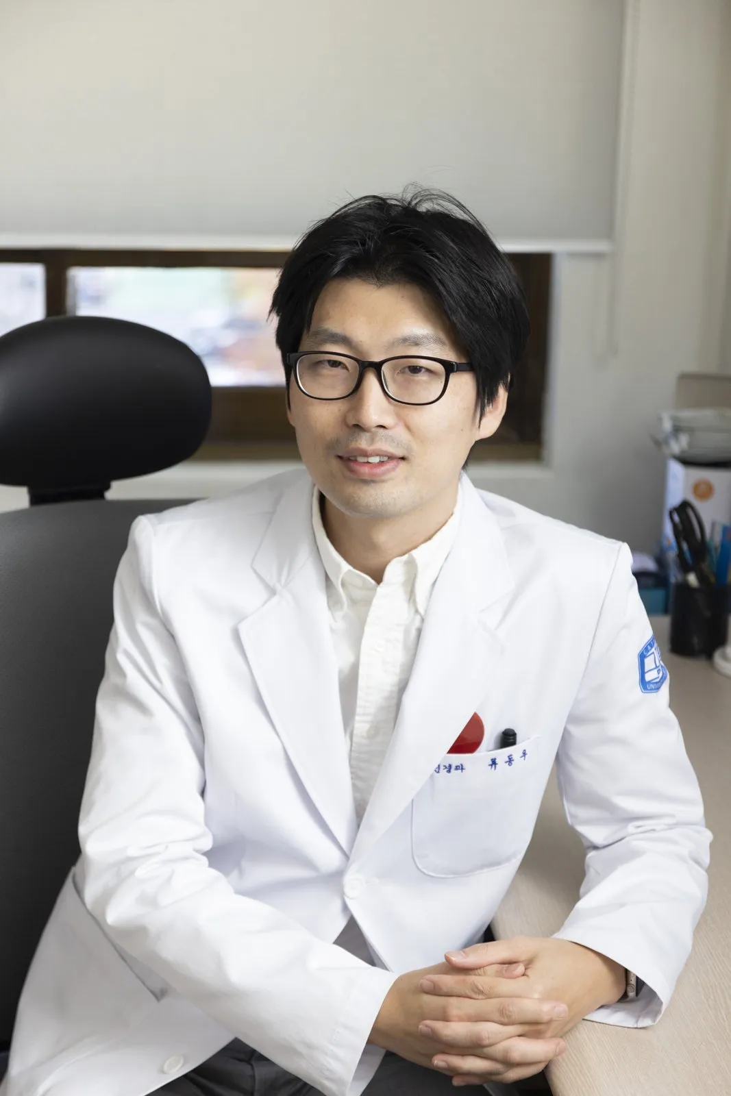 Portrait of a doctor with glasses and a white coat, seated in his office with his hands clasped