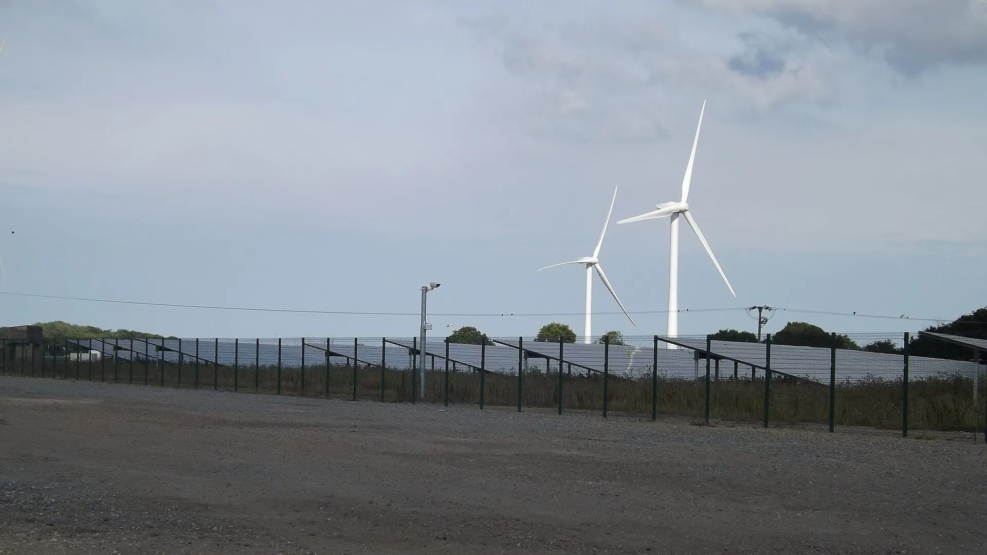 Rows of solar panels behind a fence, with two white wind turbines in the background under a cloudy sky