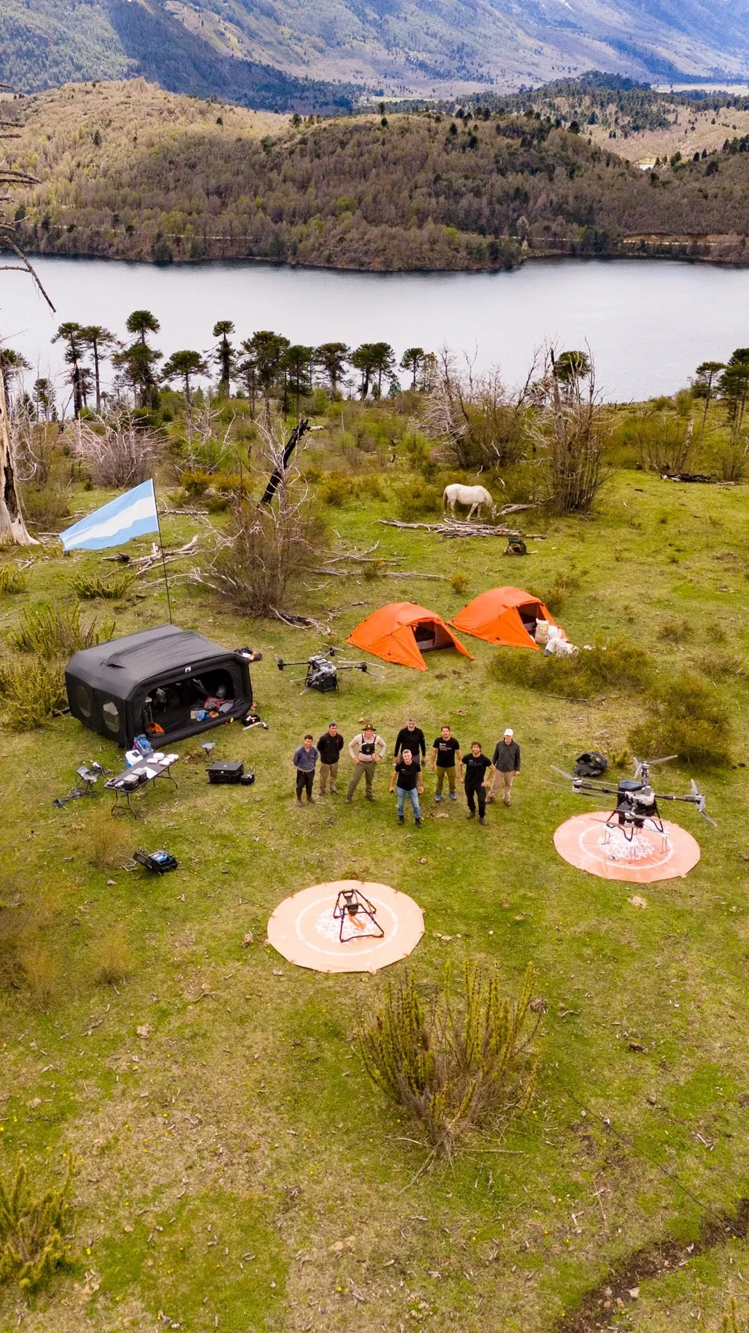 Aerial view of a camp in a meadow by a lake: two orange tents, a black shelter, two drones on landing pads, a white horse and a group of eight people looking up at the camera