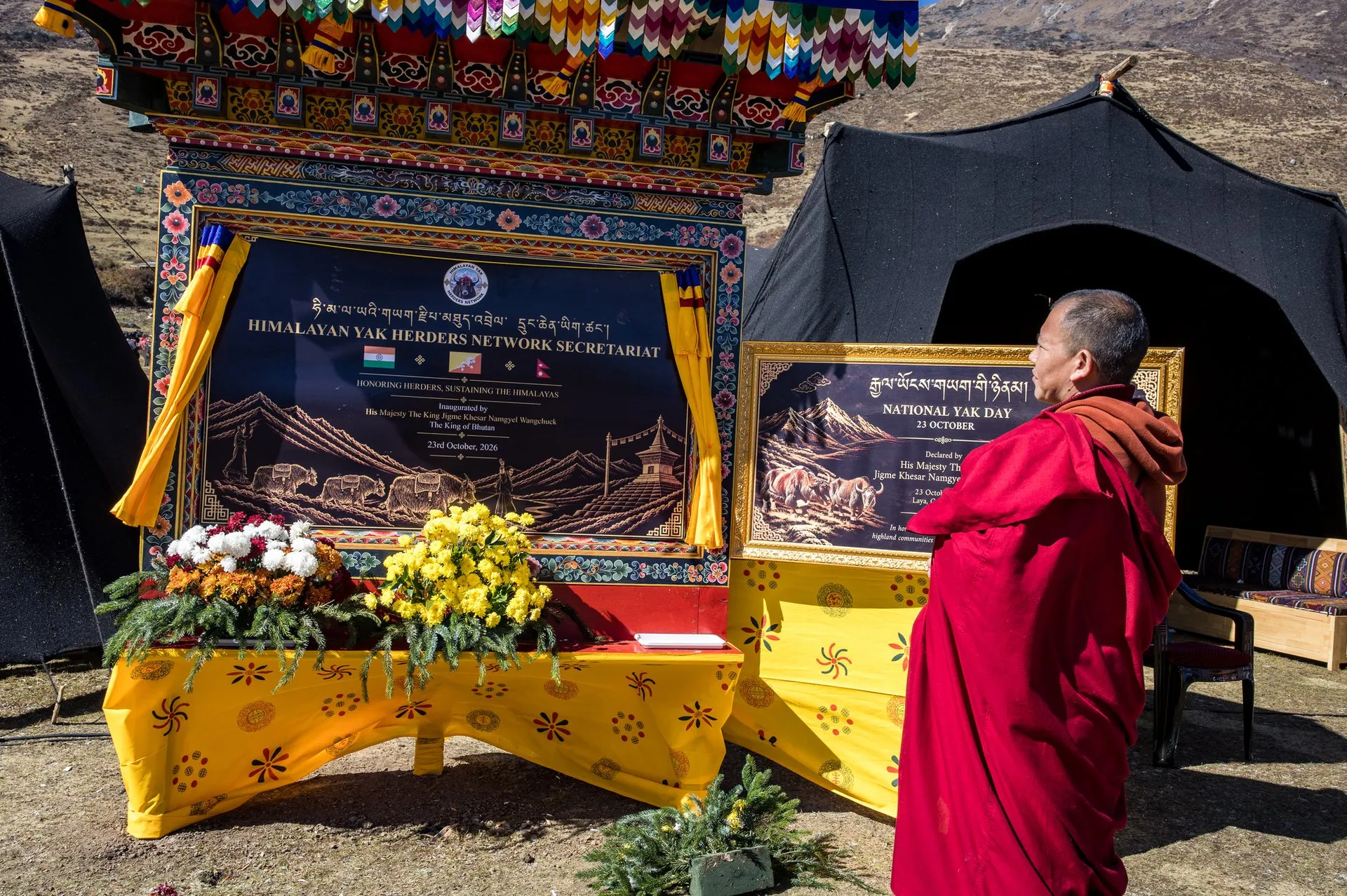 A monk in red robes stands before a painted wooden frame holding a plaque that reads "Himalayan Yak Herders Network Secretariat", with the flags of India, Bhutan and Nepal