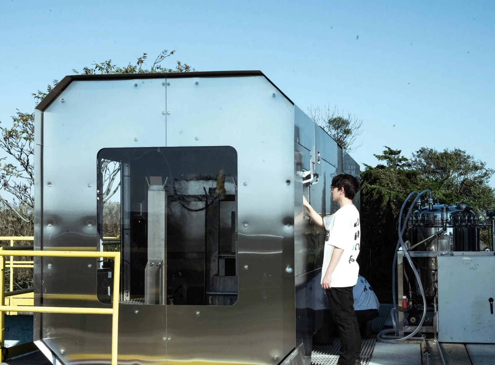 A container-sized silver metal unit with a window showing pipework, next to a person on an outdoor platform