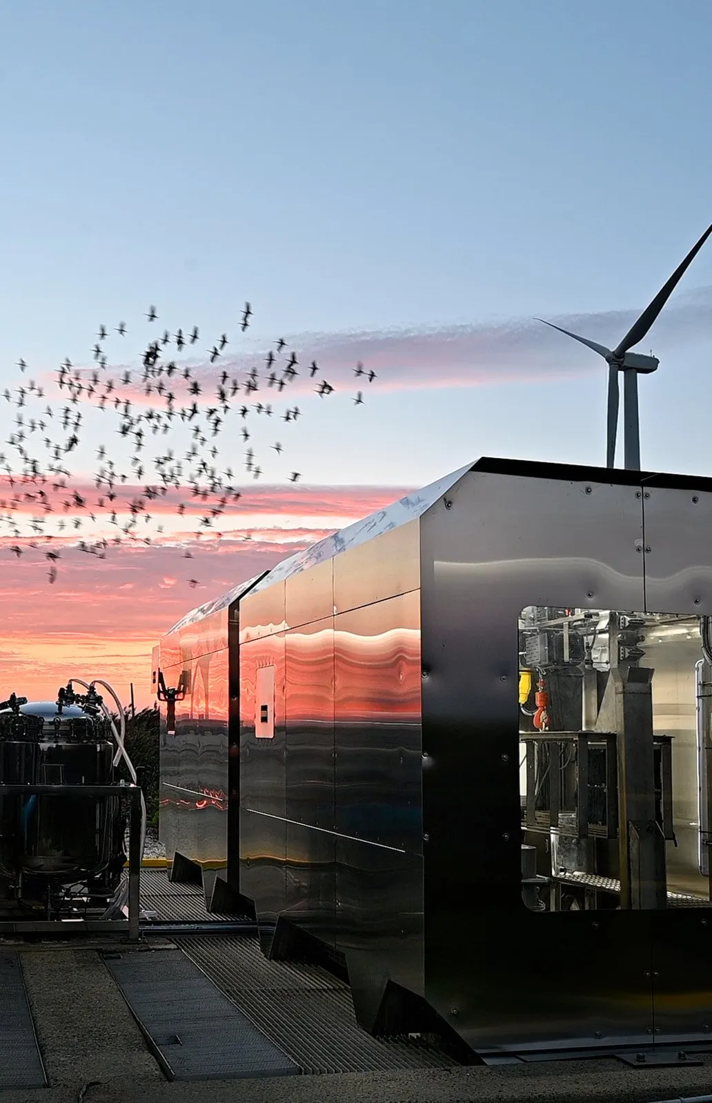 A metal unit at the treatment plant reflecting an orange sunset sky, with a flock of birds and a wind turbine behind