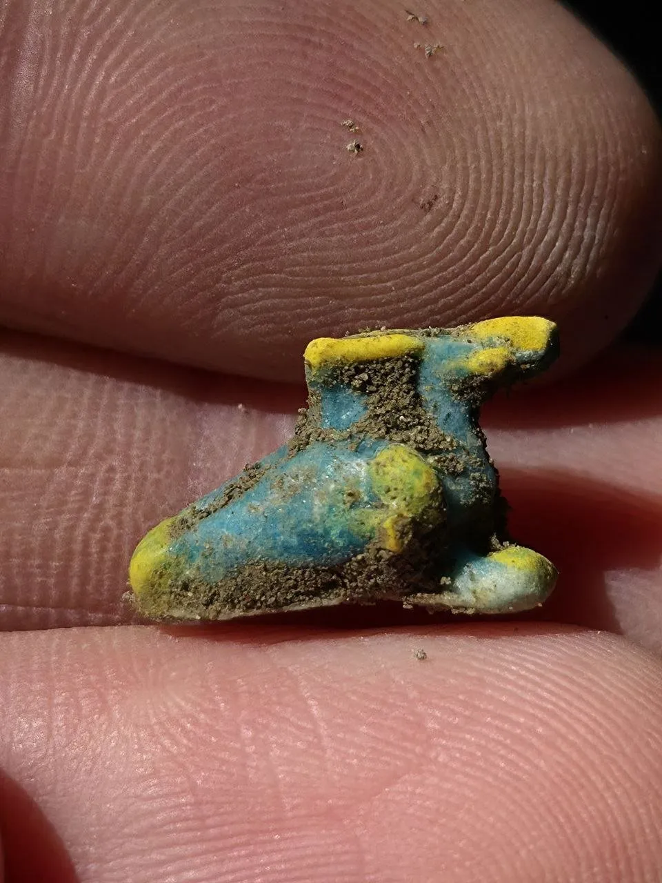 A small blue and yellow charm shaped like a winged sandal, held between two fingers