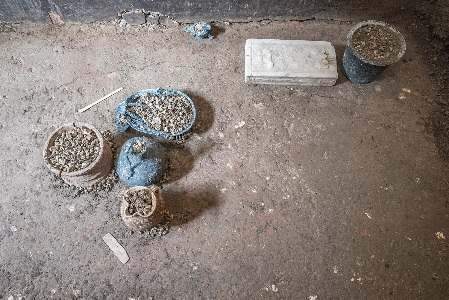 Earth-covered bronze and pottery vessels next to a white marble box on the floor of an excavation