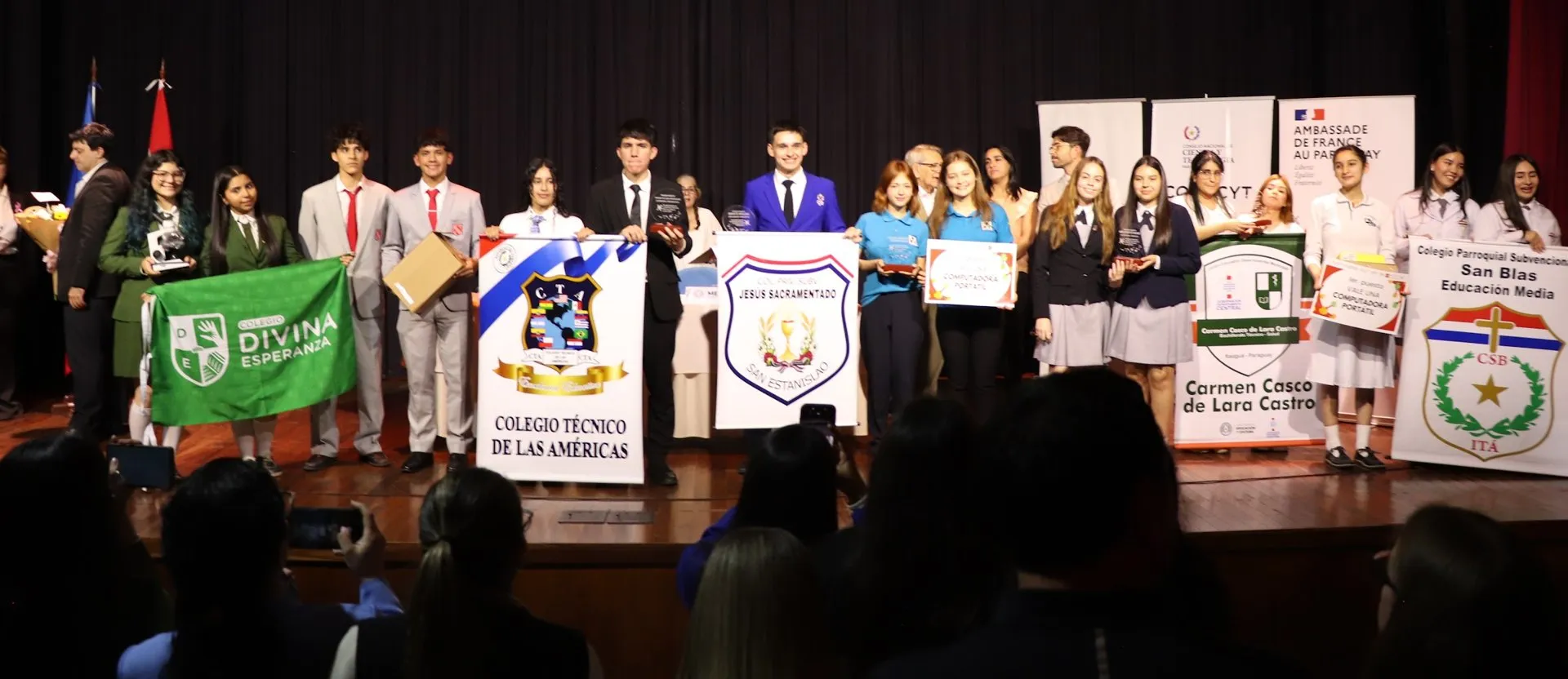 Students in different school uniforms pose on a stage with their schools' banners and their prizes