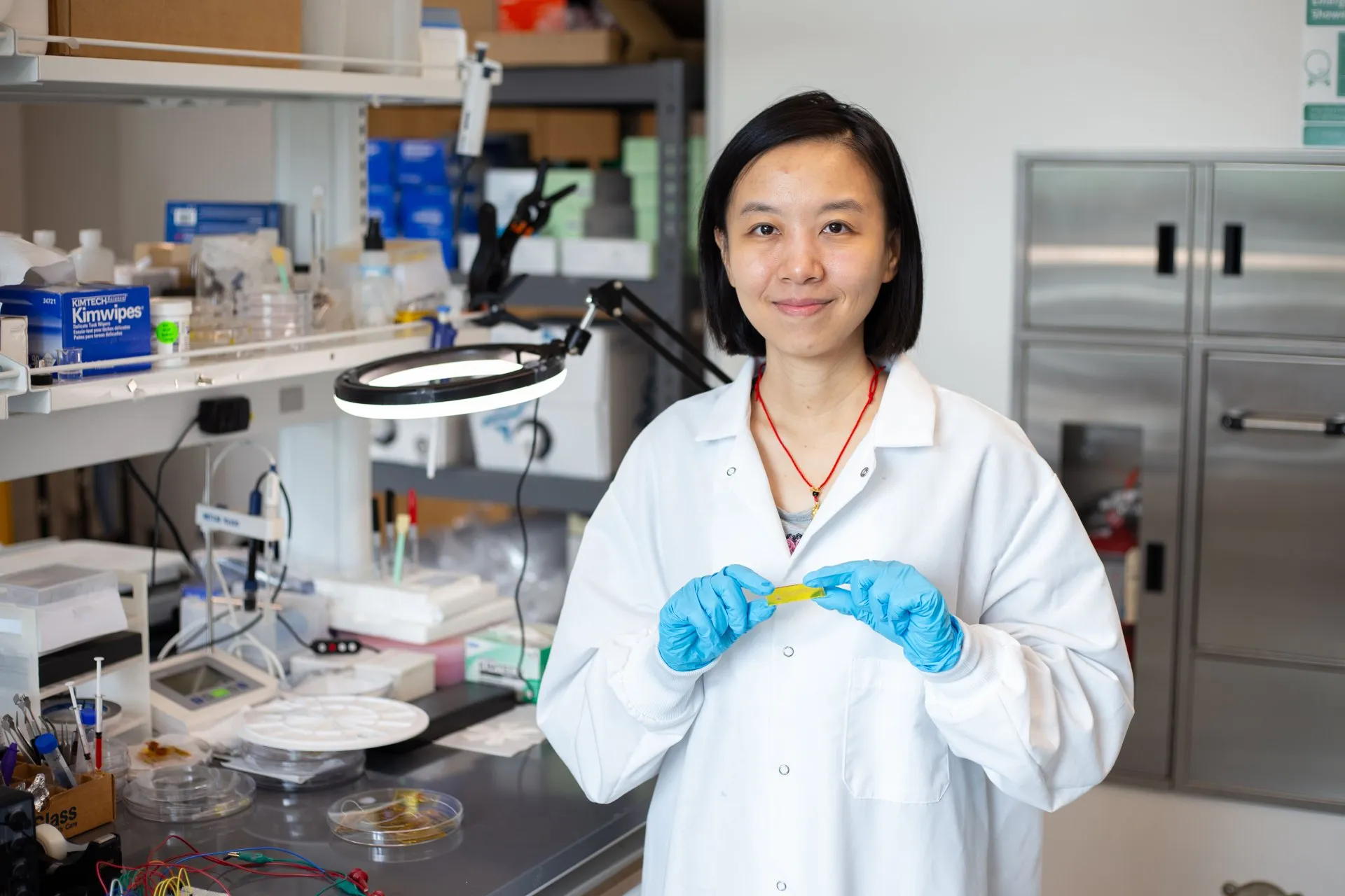 A researcher in a white lab coat and blue gloves holds a small yellow piece in a laboratory
