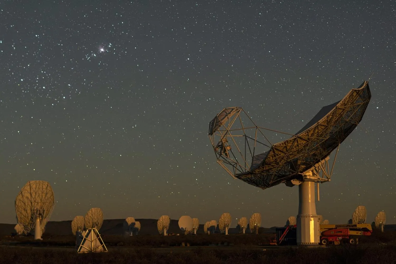A large metal-framed dish antenna lit up at night, with many smaller white dishes in the background under a starry sky