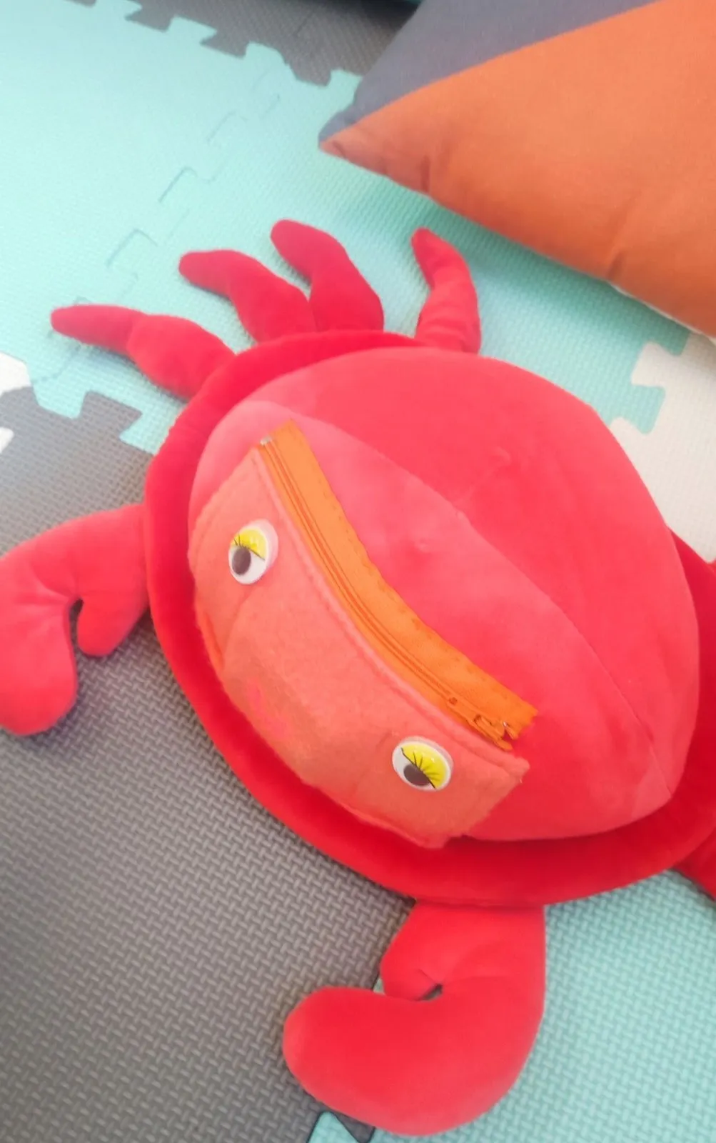 A red cuddly crab toy, with googly eyes and a zipped pouch on its shell, lying on a colourful foam mat