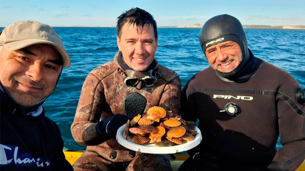 Three smiling men in a boat, two in wetsuits, hold a plate full of orange scallops with the sea and the gulf coast behind them